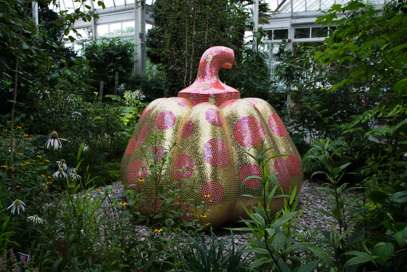 a whimsical and elegant large pumpking sculpture colored with  shiny gold and pink tiles sits amdist echineca flowers and greens in New Yorks Botanical Garden