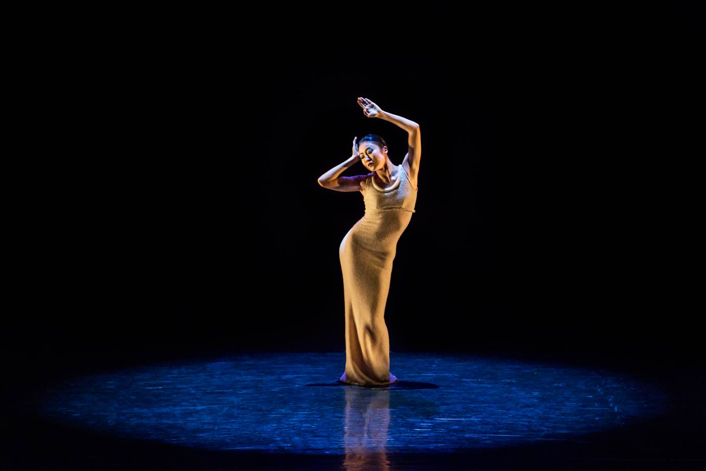 A woman in yellow snugly fit long dress, rests one hand on her forehead and lifts the other in the hair. She stands in a narrow position under a spotlight.