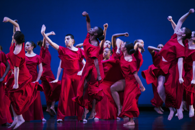 A group of young dancers in bright red shirts and long skirts lift their fists in the air.