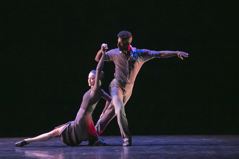 A man holds his female partner by the hand as she descends in a split to the ground. They both wear costumes in grey hues.