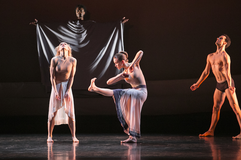 Two dancers jut their chins out to the ceiling and stand in contracted rigid poses. A dancer stands between them balancing on one leg. A dancer in a billowing cloak is in the background with arms outstretched.