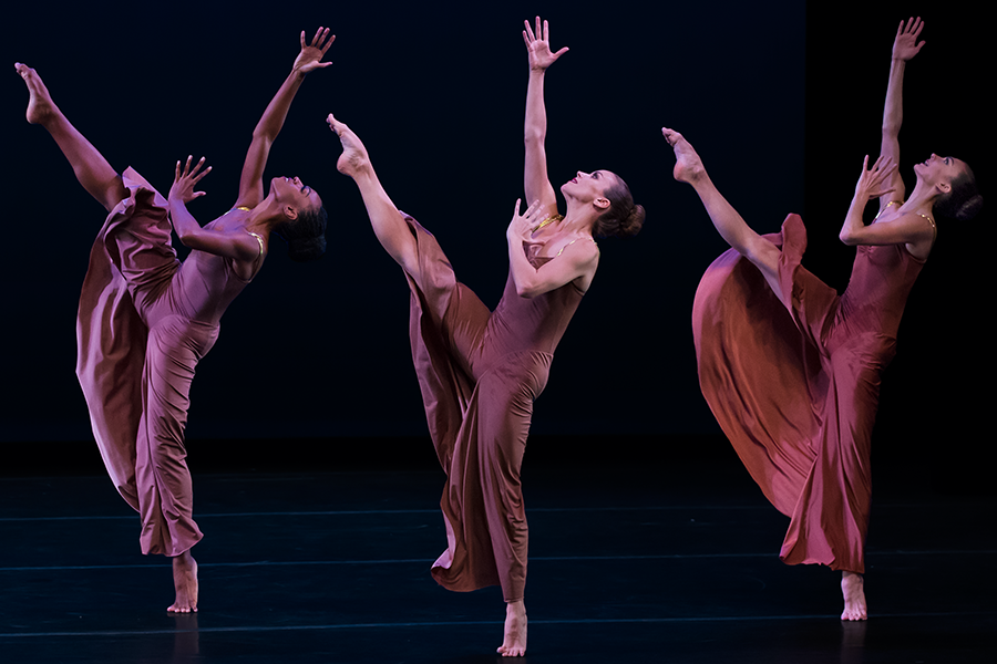 Three women facing profile, lean backwards kicking their legs high into the air. The reach to the sky.