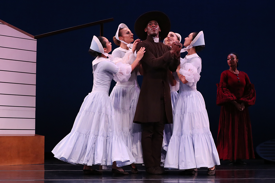 the preacher in the center clasps his hands together solemenly looking outwards as his followers dressed in tiered gowns of blue with ruffled white sleeves and dainty blue and white caps, gaze up at him adoringly, behind this scene stands the pioneer woman, a long burgundy gown, looking rooted, wise, and constant 