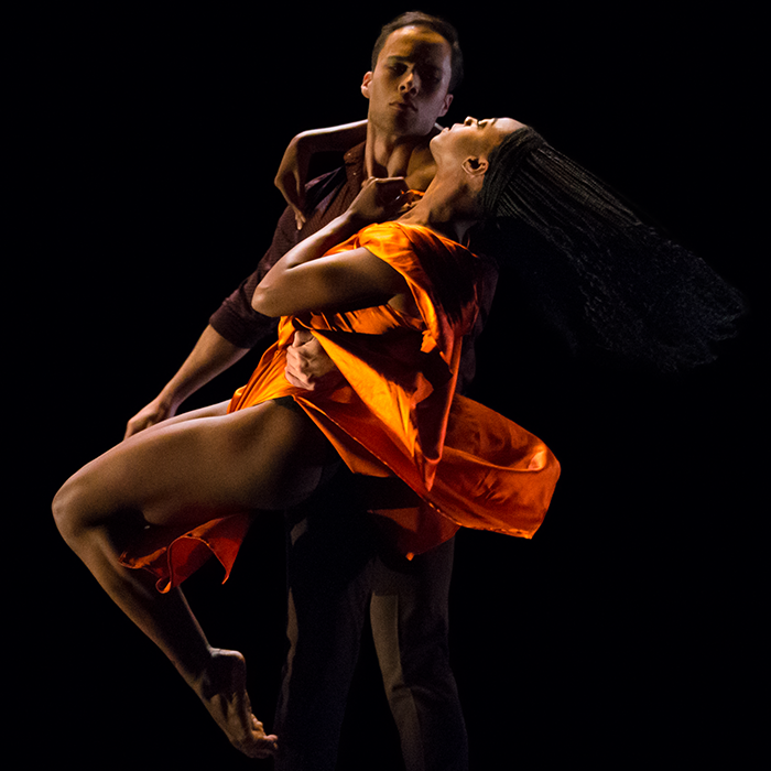 cloaked in darkness a black woman in an orange dress is swept up by a man of color. She is in profile looking up at him, her legs revealed as her dress swings to the back of her, We can only see the man's face and that he is wearing a dark shirt
