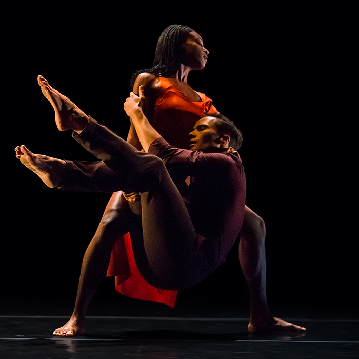 The woman in the orange dress stands in a wide plie as the man dresed in maroon desperateily clings to her.She seems serene and strong.