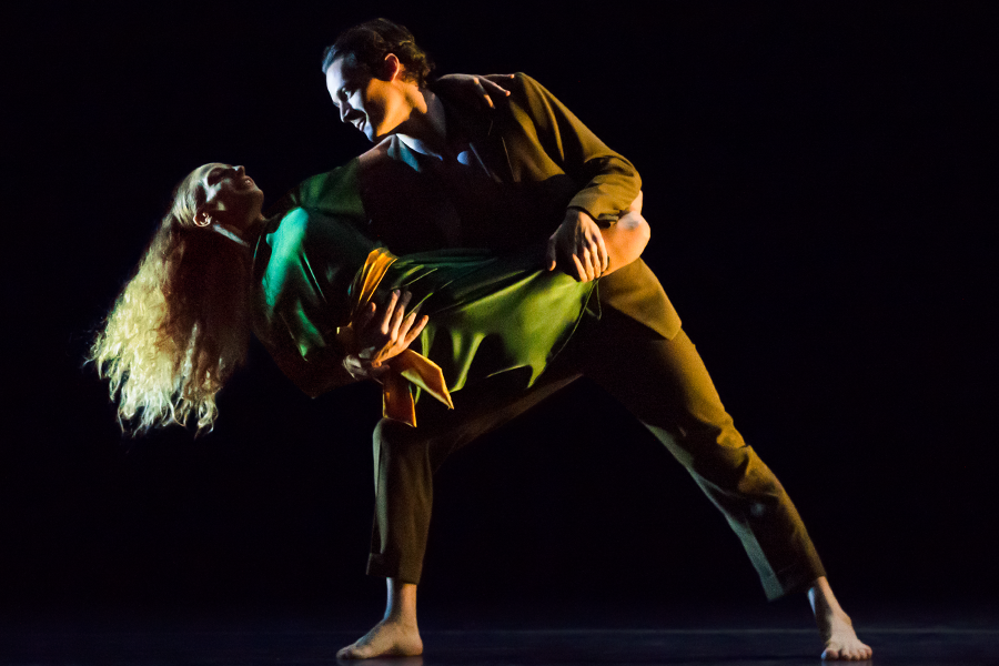 a woman in green satin dress with long blonde hair, wraps her legs around her olive suited partner as he carries her. He stands in a lunge and though her chest faces away from him she twists her upper body so she can look into his eyes.