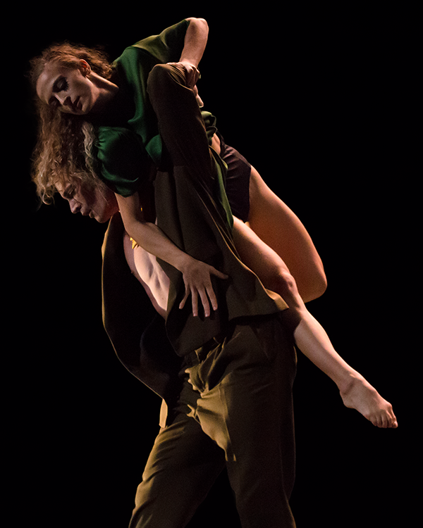 The blonde woman is being carried on the back of her olive-suited partner. Her dress has slide up revealing brown satin underwear. We notice her suited partner is shirtless.