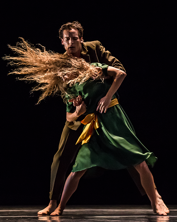 the olive suited partner man with brown hair grabs the upper torso of his female partner in front of him. Her blonde her swings to the left almost perpendicular to the floor.For the first time we notic she has a yellow belt.