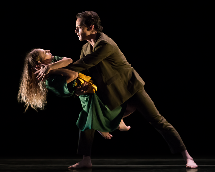 a white man with brown hair leans into a lunge facing his female partner, her legs are wrapped around his bent leg and she grabs his hand and peers into his eyes. Both look at one another lovingly.