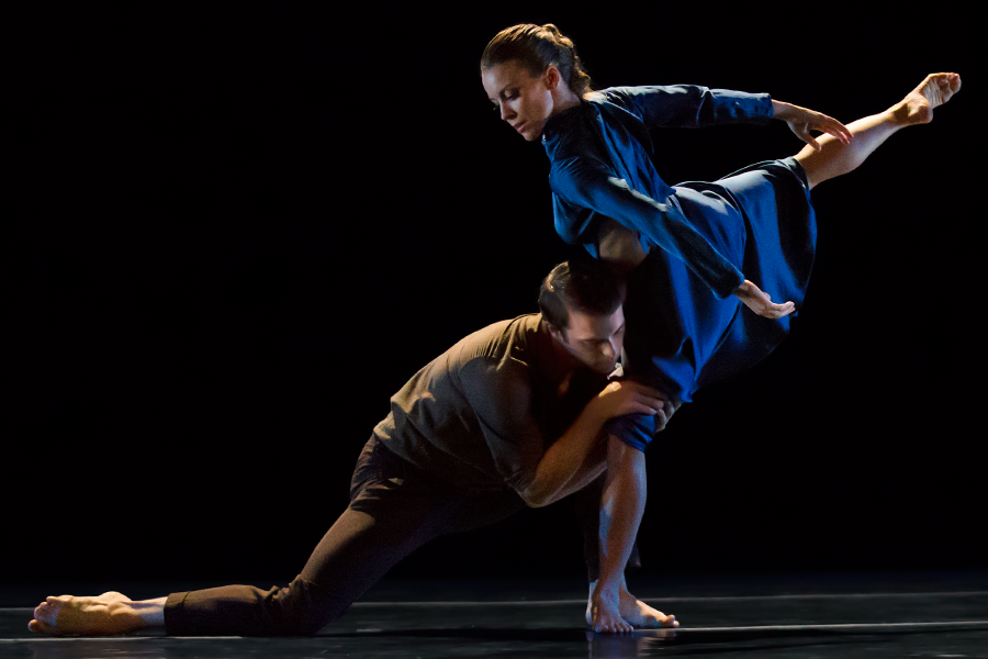 a woman wearing a blue satin long sleeve dress demonstrates a beautiful arabesque as she looks down at her male partner. He is in a low lunge holding on to the knee of her standing leg quite tenderely.