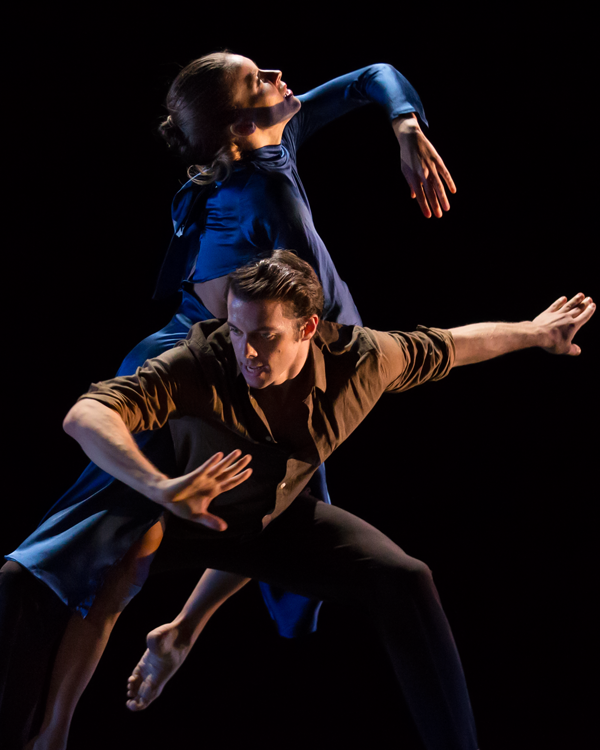 the woman in blue rides on the back of her partner looking upwards past her extended bent elbow..The man faces the opposite direction looking downward to the floor.