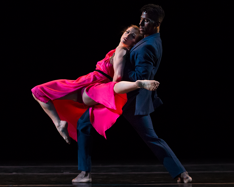 the woman in red lies across her partners chest and looks out to the audience. One leg is extended in the air, towards the audience, the other is extended in front of her with her knee bent. 