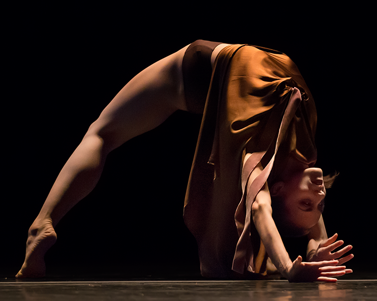 the solo woman dressed in brown assumes a yoga wheel position as part of her dance.her lige and upper body are lit, as well as her face and fingers. She almos looks like a spider.