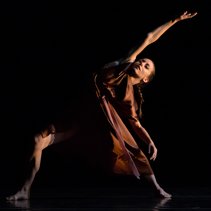 The solo dancer's knee peeks out from her brown satin dress as her body arches away from it. she is fimly planted in the ground one arm extends up ward the other is relaxed dowards the floor. Her eyes are closed.