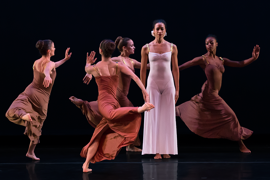 the woman in white stands still and solemn as a chorus of women with large buns dance around her 