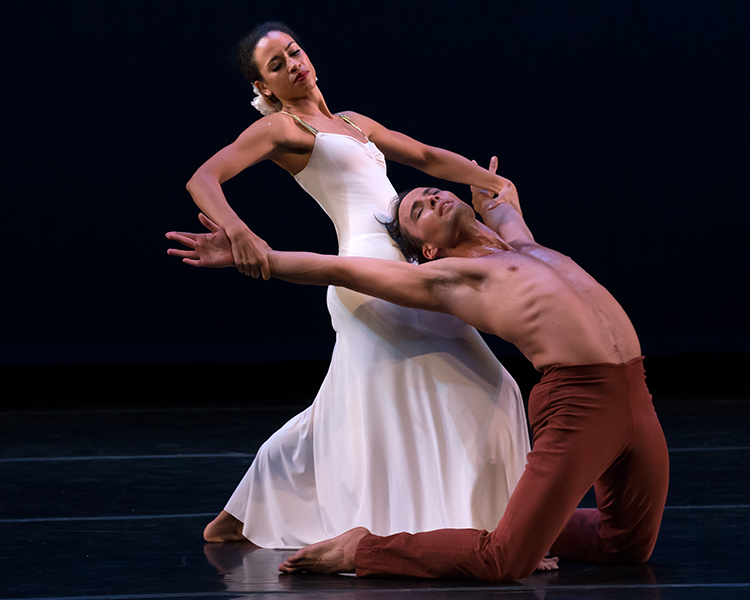 the woman in white grabs her lovers spread arms as he arches backward from his knees in a moment of passion. his head appears to be leaning on her stomach