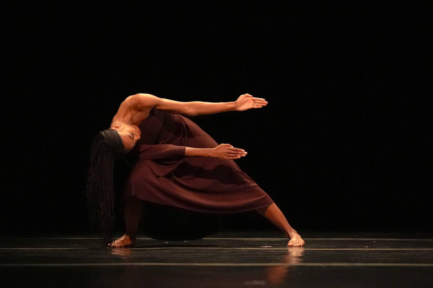 A woman dressed in an off-one-shoulder long brown dress, bends sideways with her head near her waist, thrusting both arms horizontal to her left. The left palm is up and the right palm is down.