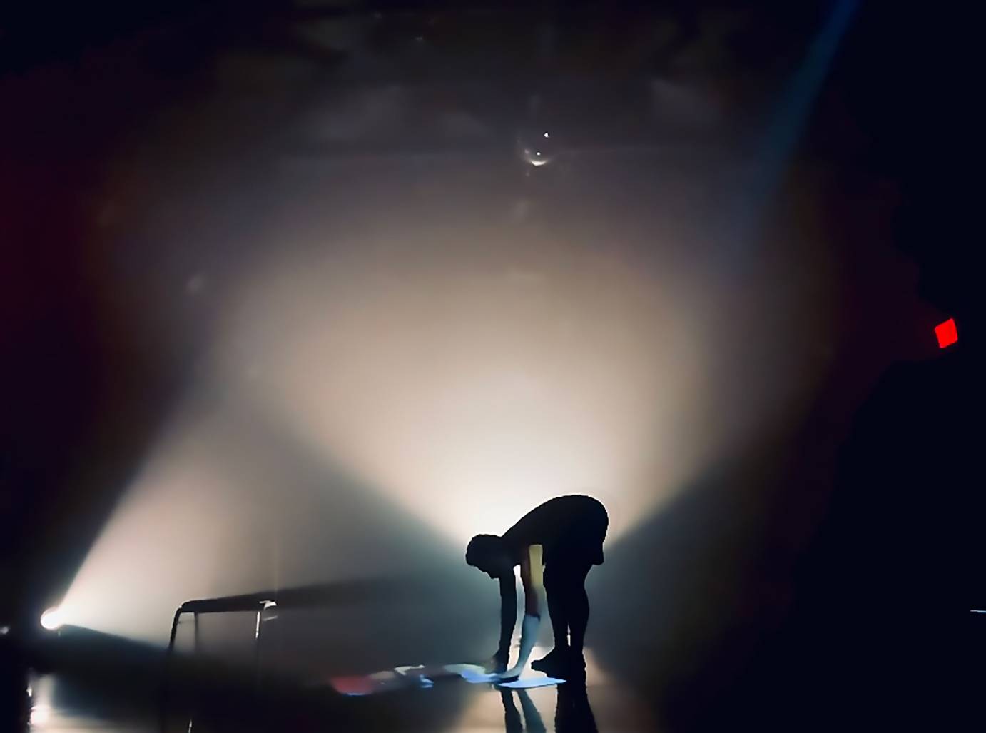 A figure, bent forward, bathed in white light against a black background, studies a paper on the stage.