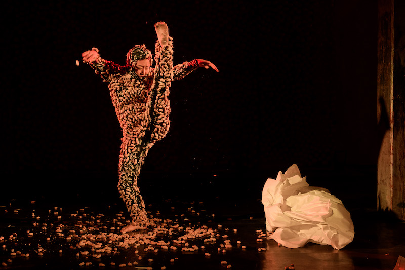 An artist in a onesie covered in peanuts kicks her foot. Peanuts litter the floor around her.