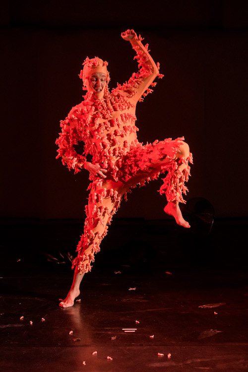 A woman in a bright orange unitard with tiny plastic babies attached.