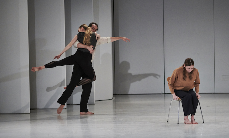 One dancer holds another while a third sits in a chair wearing a camel-colored turtleneck.