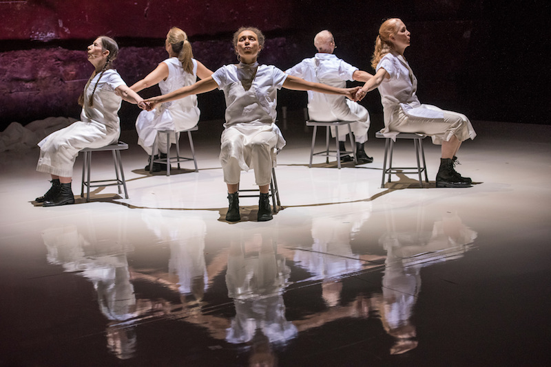 Five dances sit on low stools in a circle facing out. They hold hands and sing.