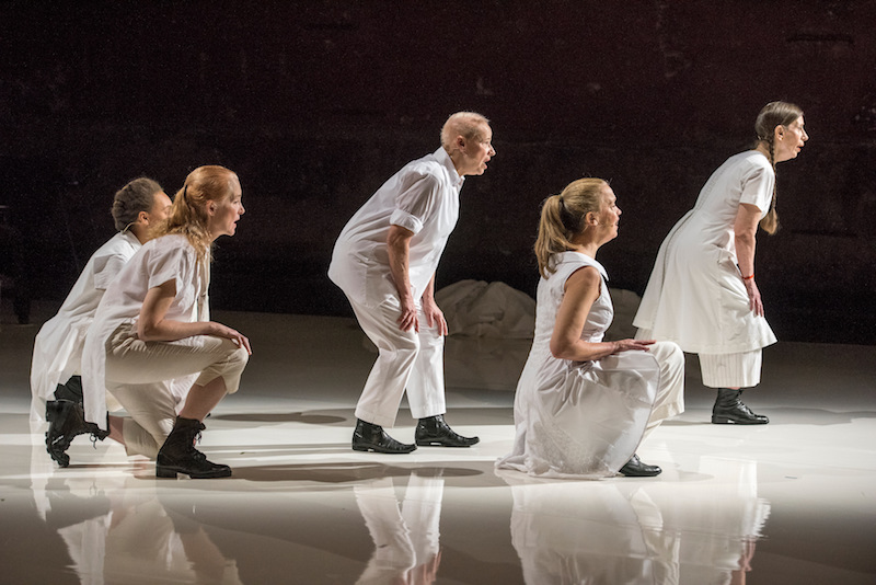 Five performers crouch at various levels and sing. They wear white and we see them in side profile.