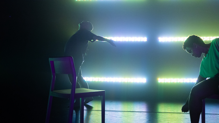 Under a green cloudy light a man in street clothes arches his back upward a chair is in the foreground, next to it is another chair where a black woman in a green top sits on another chair and reaches toward the floor.