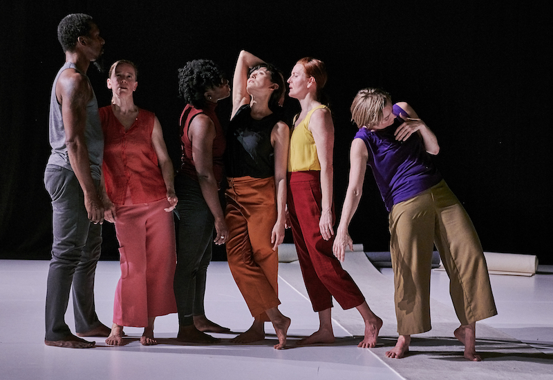 A group of dancers in bright color tee shirts and chimps stand in a cascading line. Some lean on one another. Some face away from the camera.