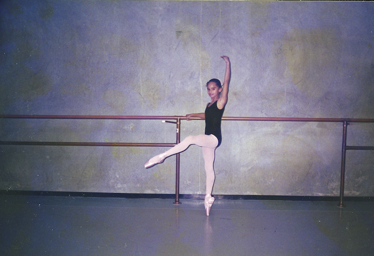 a little Misty Copeland at the barre in a dance studio with gray walls... this is her first time in pointe shoes and she is elgantly balancing on one leg.