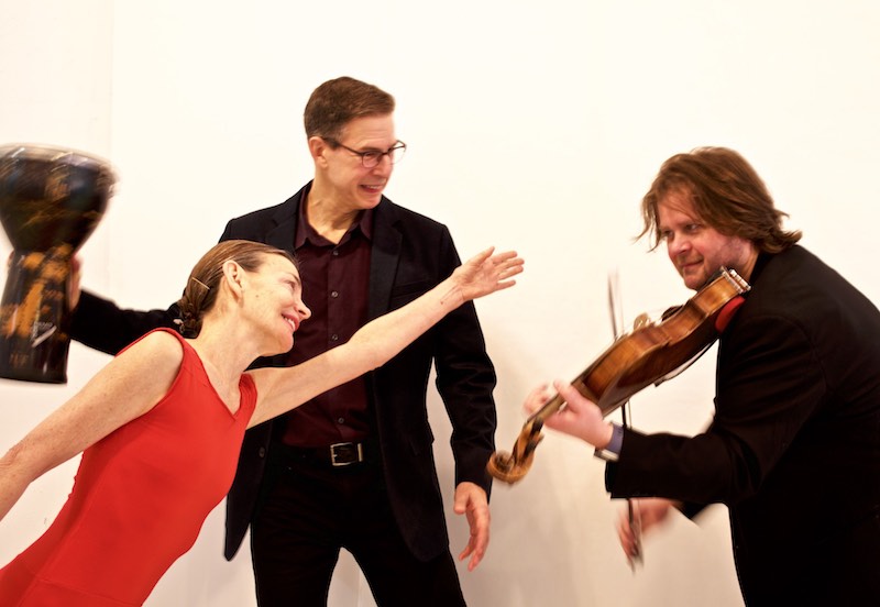 Molissa Fenley in a red shirt outstretches her arm toward violist Ralph Farris. Drummer Frank Cassara smiles. 