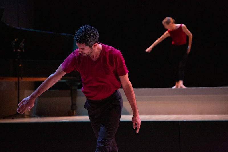 Two dancers in burgundy tops and black bottoms create a simple but riveting architectural shape by simply standing in a low hover and extending one arm to the side. The fact that each dancer stands on a different level of the stage adds to the spatial interest