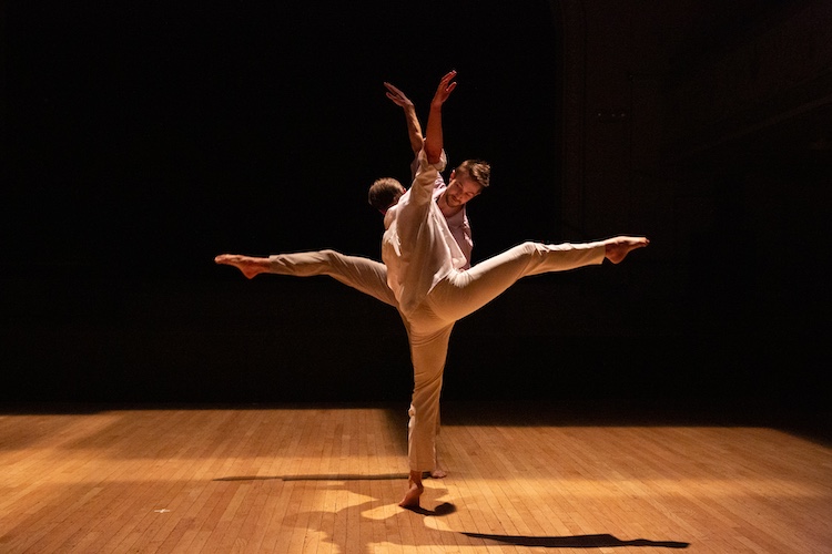 two dancers in white jump suits balance in a tilt with one leg and one arm in the air . Facing eachother so close together the form one unified shape.