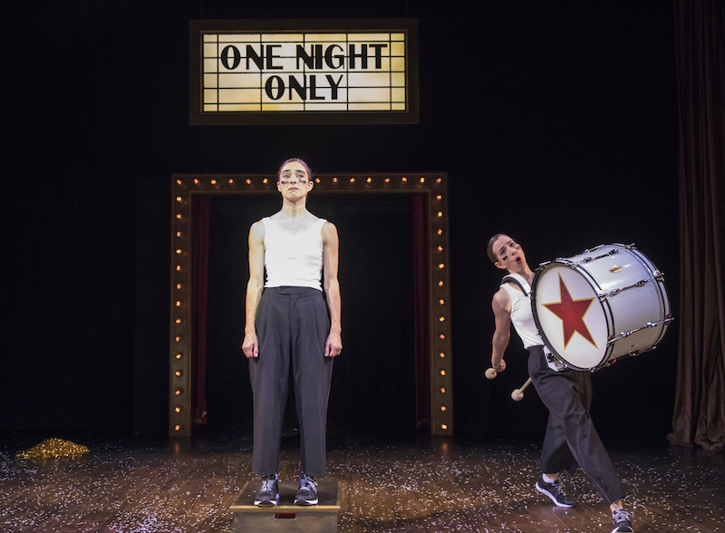 Monica Bill Barnes stands on a box looking resolute. She and Anna Bass wear black trousers and white tank tops. Anna Bass wears a marching band bass drum. Confetti litters the floor.