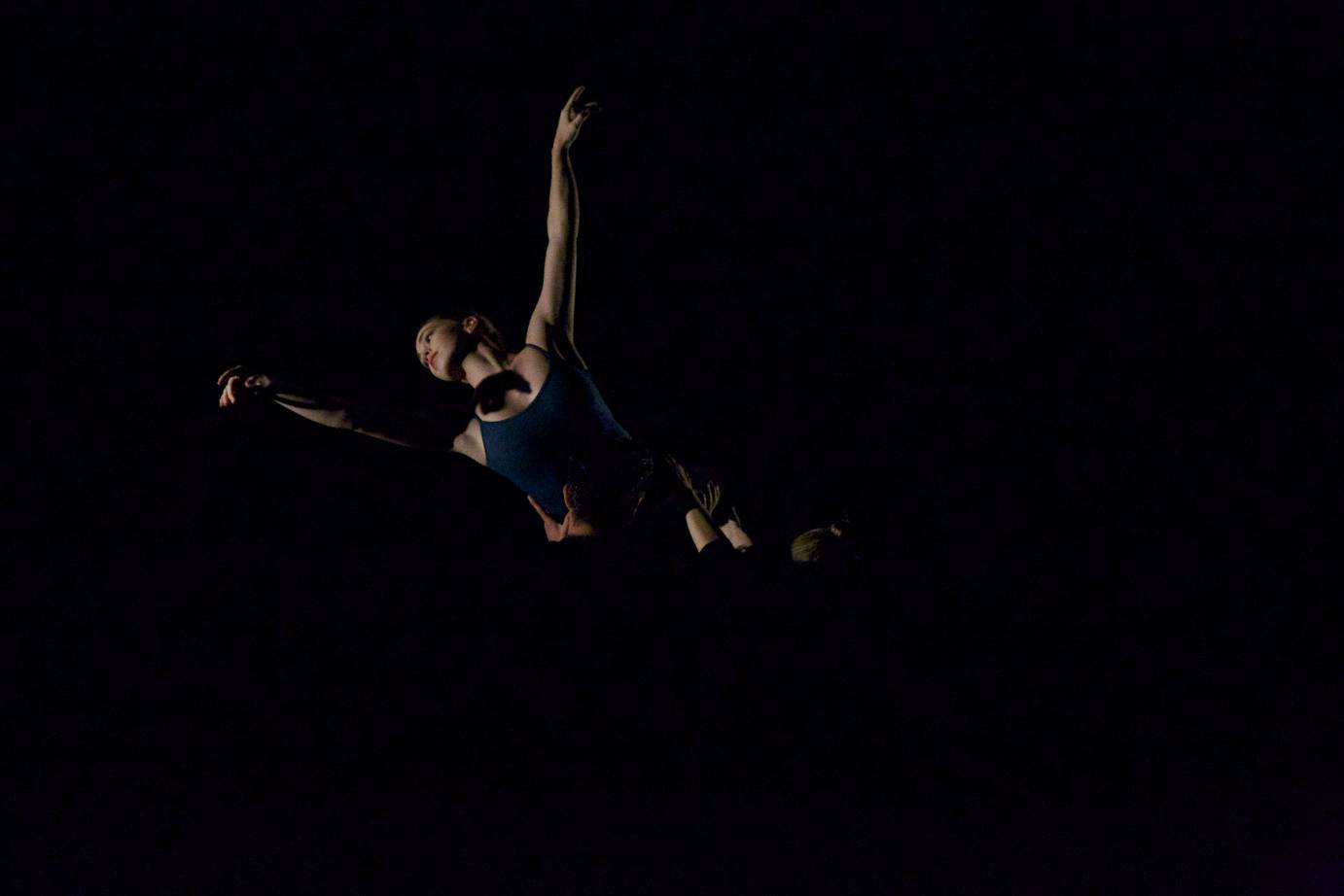 In partial darkness a women is lifted by a group of dancers in the air so that she looks like she's floating