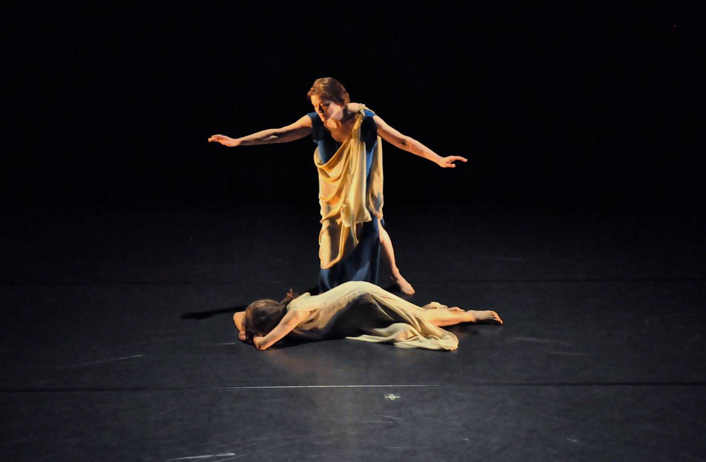 One woman in Grecian dress stands over another who is prone on the floor