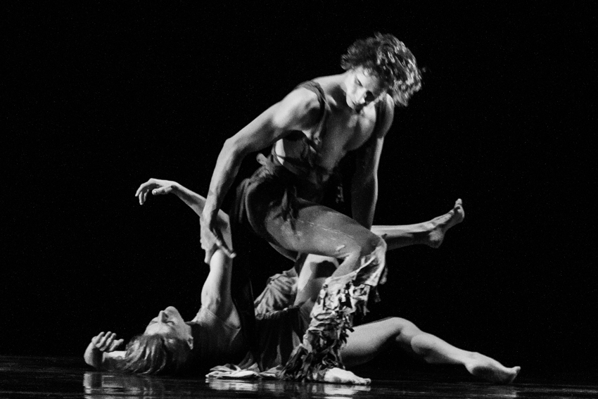 A woman lies on the ground. Her right arm reaches above her. A man in a costume that has fringe on the pant legs steps over her. Her grabs her forearm and one of her legs.