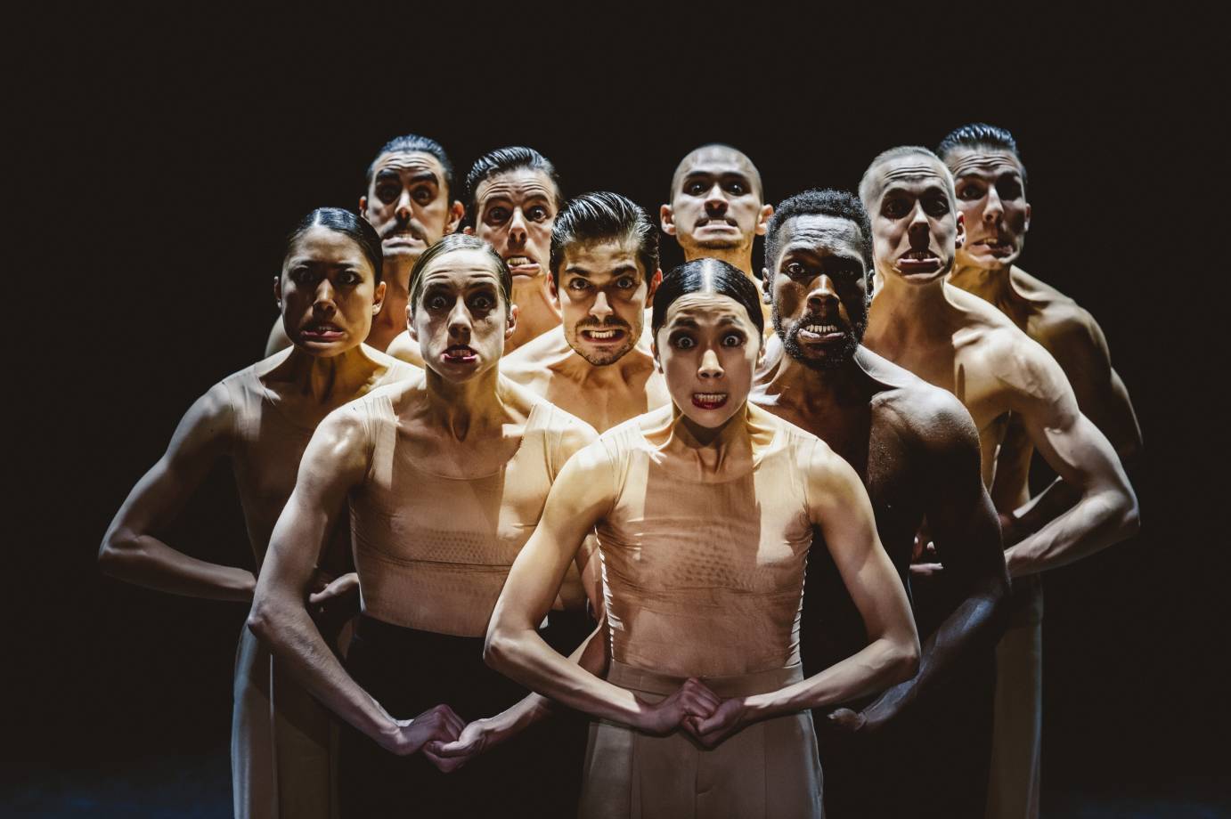 A clump of dancers clasp their hands while grimacing