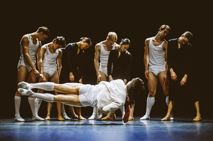 A group of dancers stand behind a man who is balancing on one arm