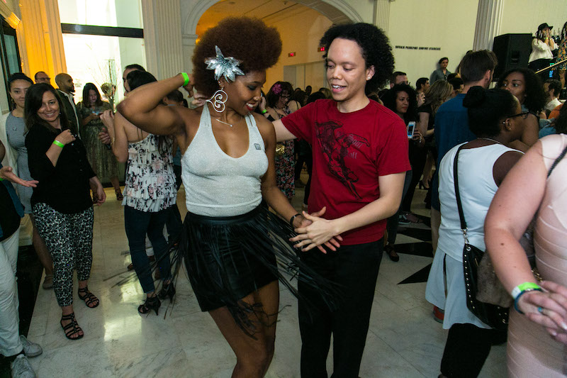 A couple dance the salsa. They stand side-by-side and hand-in-hand. The woman wears a black skirt with fringe that fans out as she moves.