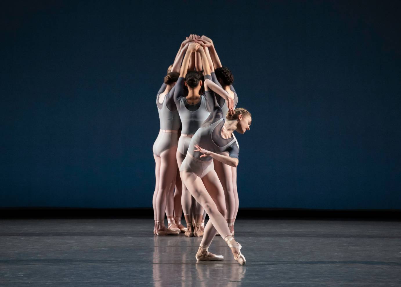 Women, with their arms extended, form a clump while another woman dances in front