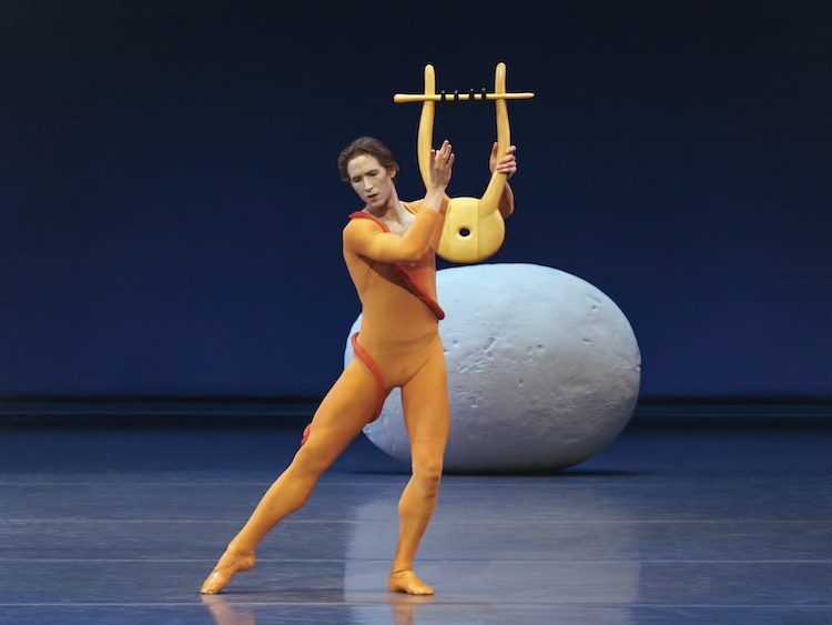 Joseph Gorden in orange plays Orpheus... standing on one bent leg with the other extended side he plays his lyre...