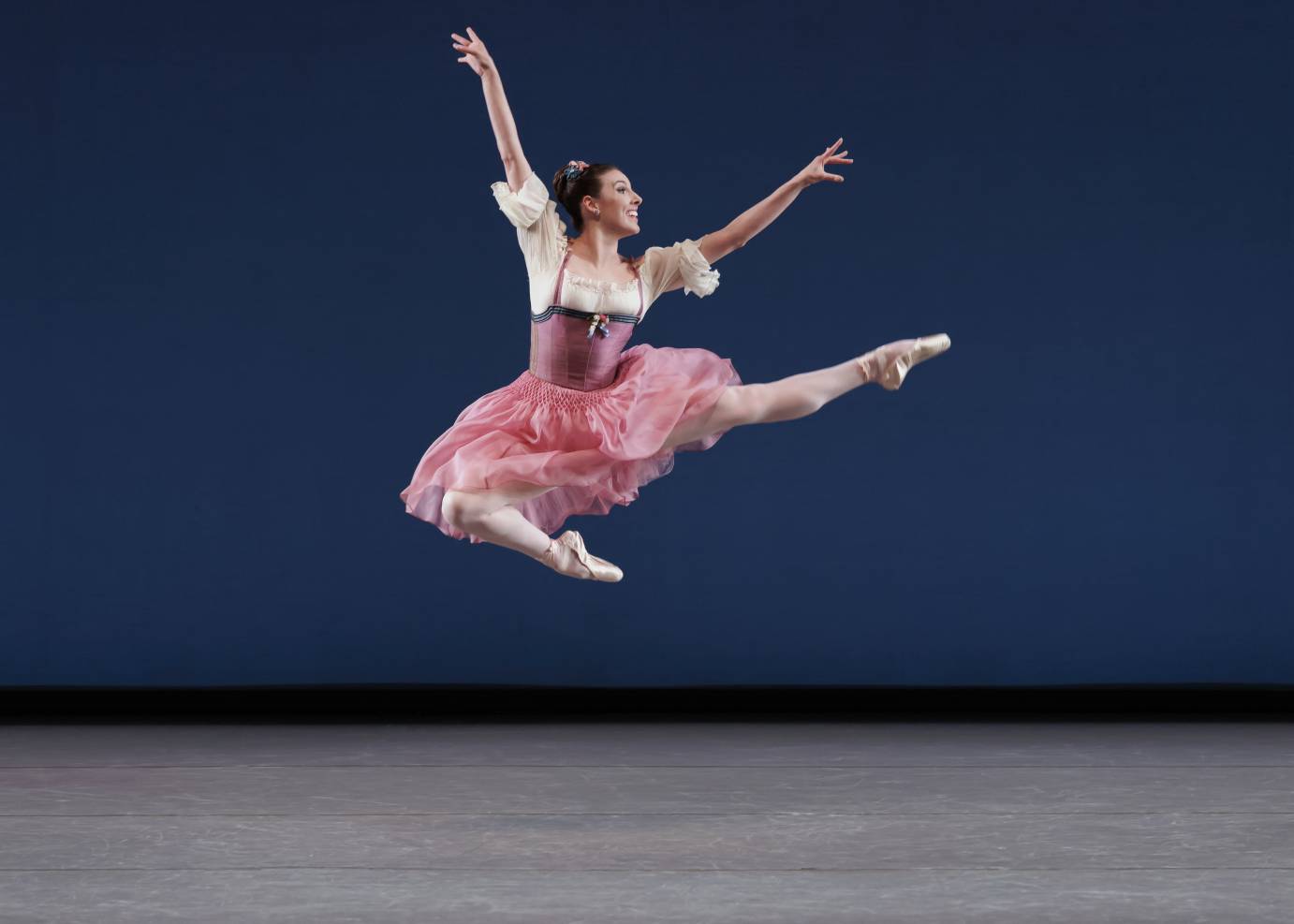 A ballerina with a wide smile, jumps high facing the diagonal, with one leg folded and the other above horizontal. Both arms are high in the air. She is dressed in a pinkish tutu, dirndle and white blouse with fluffy sleeves.