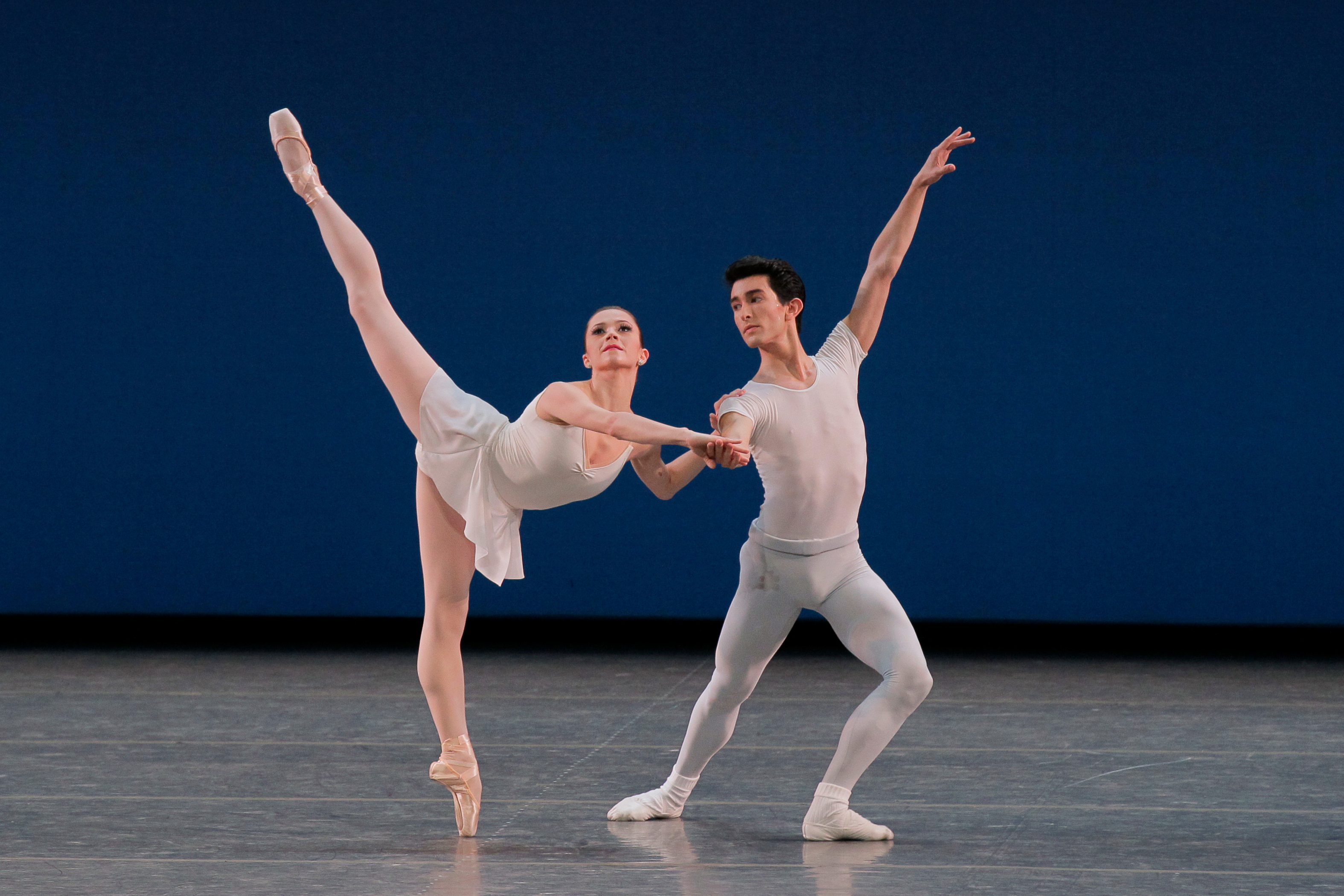 A man and woman couple, the woman on point, she lifting her working leg skyward and his left arm reaching, as well. His left leg lunges. Her face tilts up whle his looks over his shoulder and slightly down.