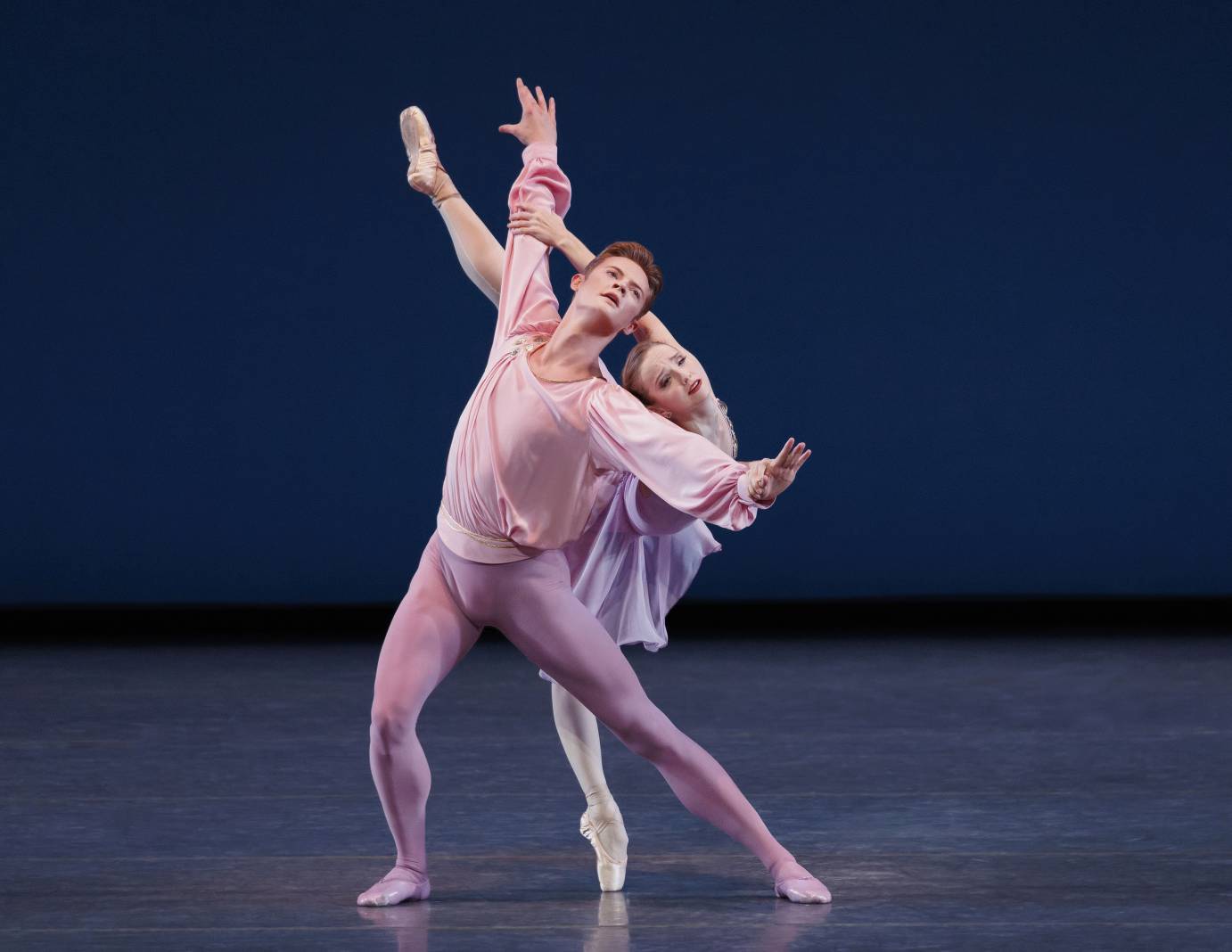 A woman and man couple dressed in light pinks and purples, backs to one another and faces full of concern as the ballerina in arabesque holds onto his outstretched arms.