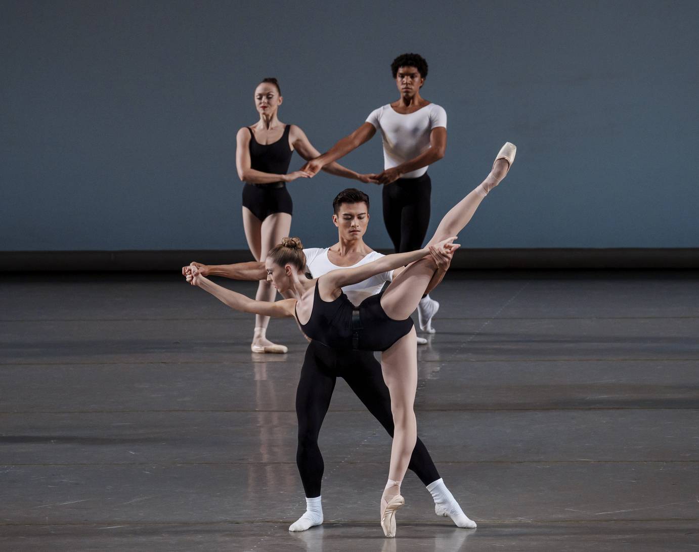 Two woman and man couples, the women dressed in black leotards, the men in white T-shirts and black tights, are positioned one couple in front of the other. Downstage, the woman, on pointe, in a deep arabesque, is held by her hands by her partner. The upstage partner, stands with one low leg behind the front standing leg, arms crossed low and hands held.  