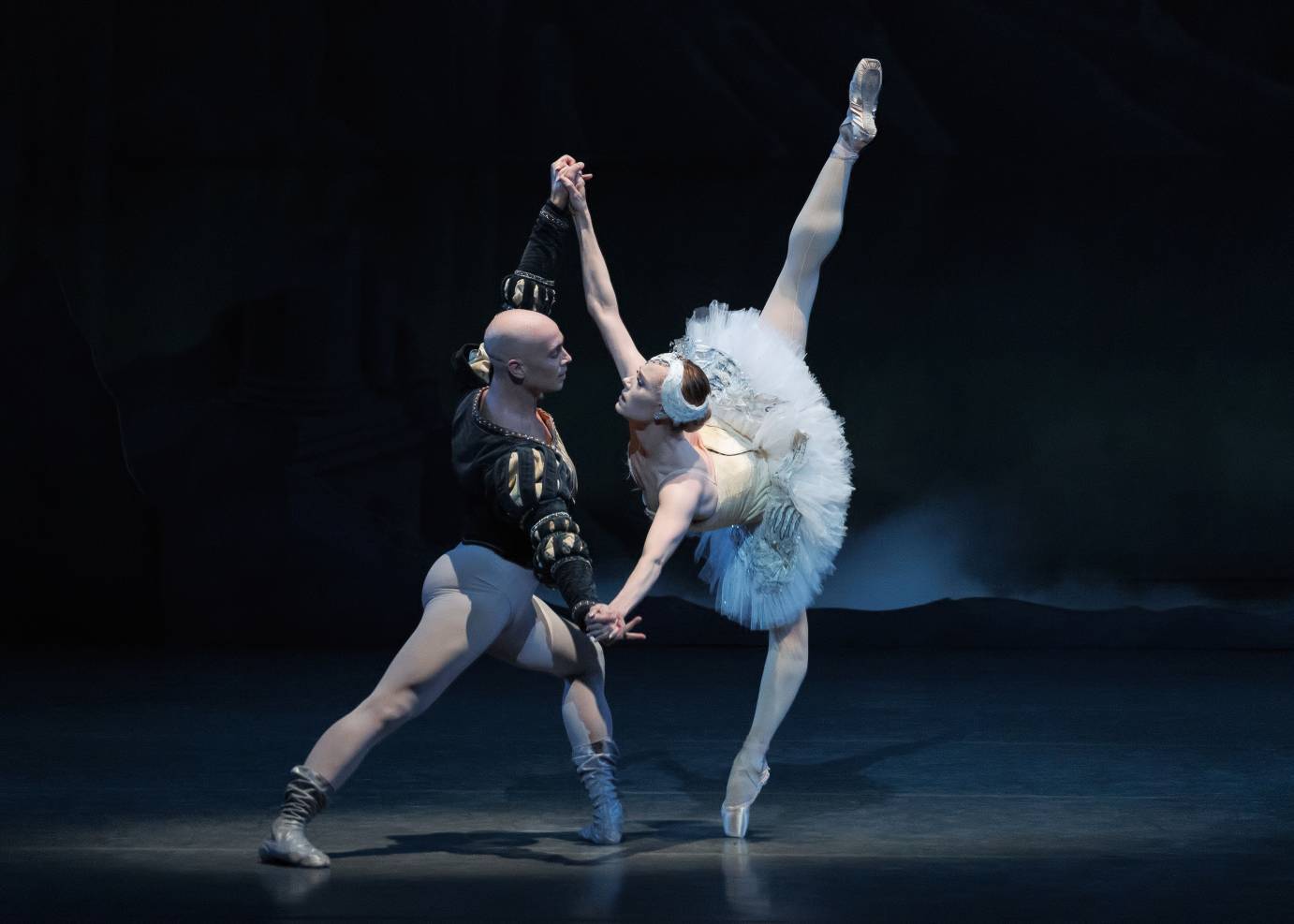 A woman and man couple face one another looking into each others' eyes, hands clasped on outstretched diagonally-placed arms, while she is in arabesque and he stands in a lunge.