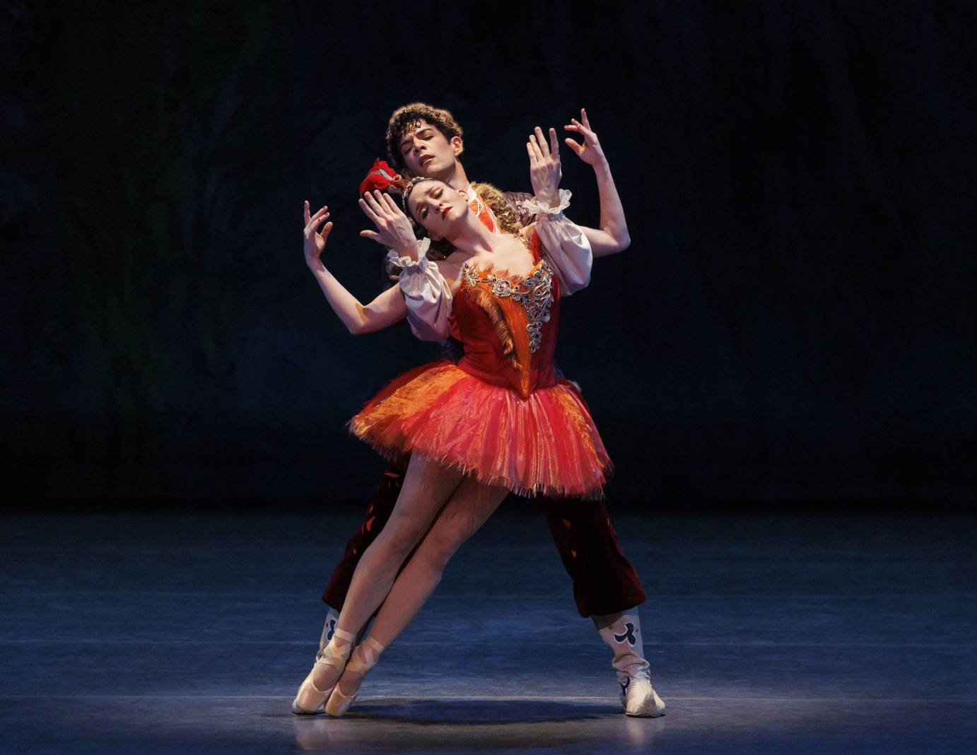A danseur and ballerina entwine arms (bent elbows fingers skyward) as she leans on him. She is costumed in a bedecked red tutu and headdress while he is in black tights. Their eyes are downcast.