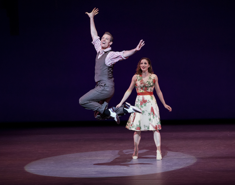 A man in a vest, wing-tipped shoes and slacks jumps into the air. A woman in a flowered dress grins behind him.