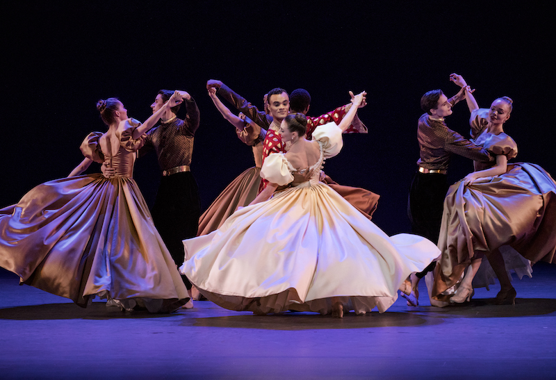 Dancers in long satin ball gowns waltz with men in silk shirts and slacks